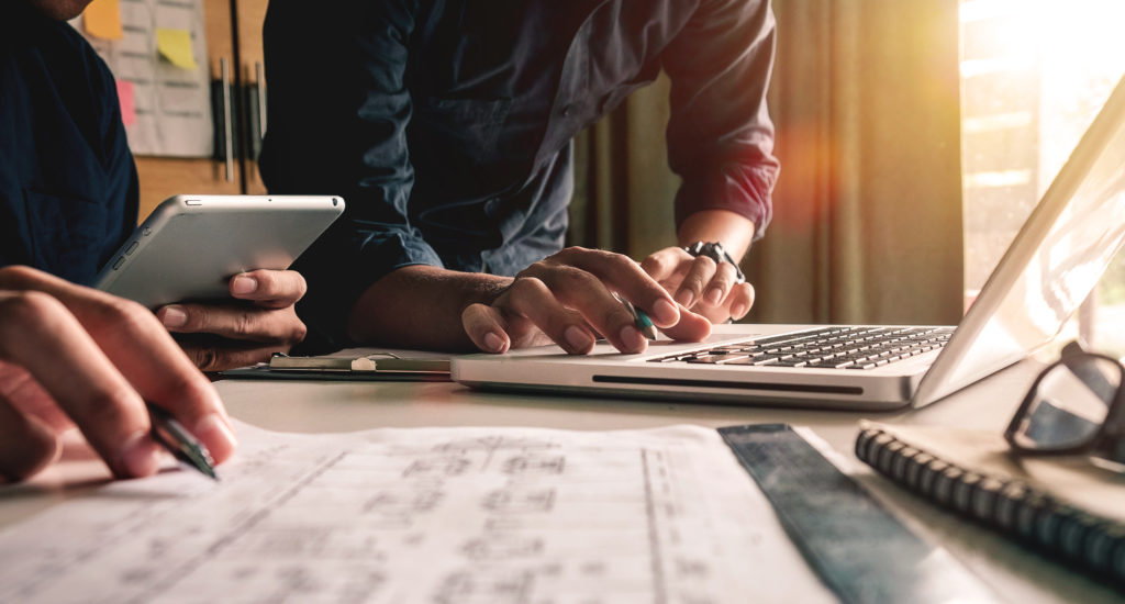 Two professionals working together on a laptop and tablet to work together on a project. The picture shows only the table and the hands of the two people. 